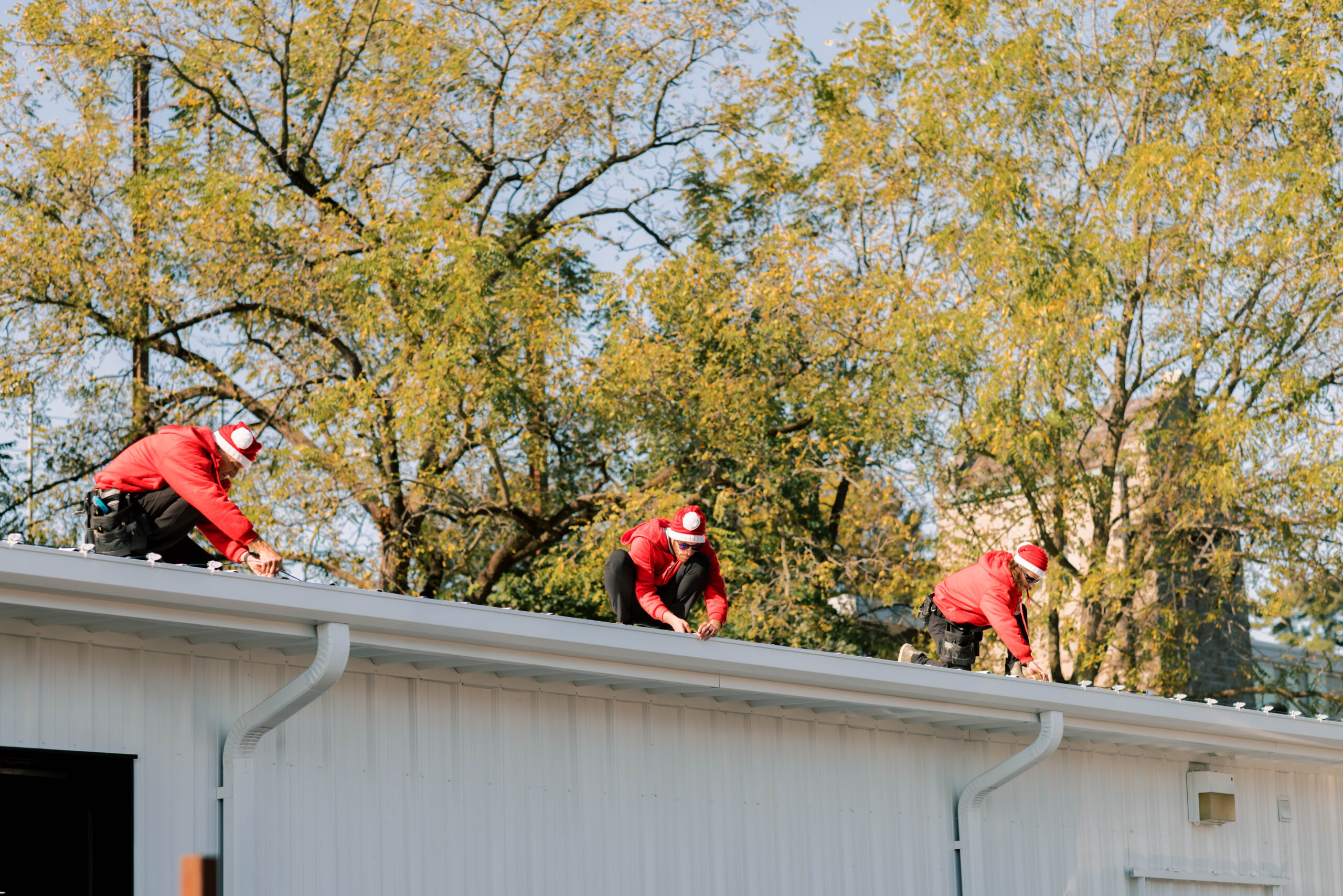 Three men dressed in red install Christmas lights while wearing Santa hats